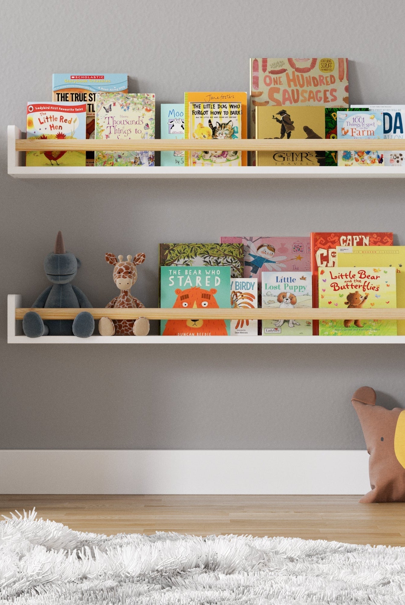 A cozy children's reading nook with two white wall-mounted bookshelves filled with colorful picture books. A plush bear pillow rests on the floor beside a fluffy rug, with a small stuffed animal sitting on the lower shelf.