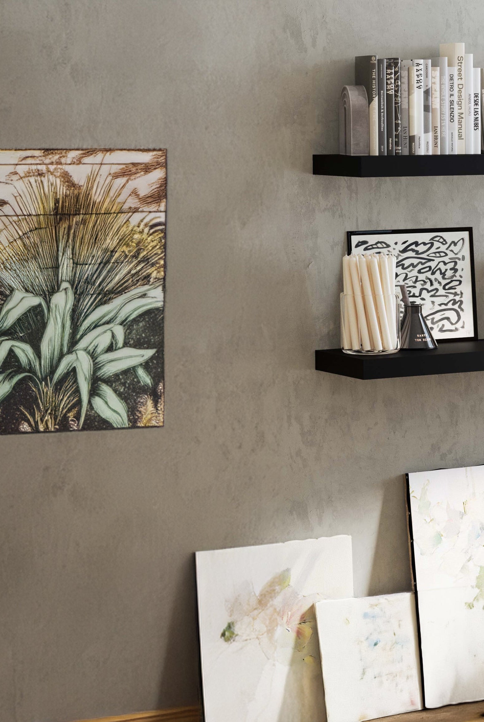 A modern space with two black floating shelves with invisible bracket mounted on a textured gray wall. The shelves hold books and decorative items. Below, framed artwork leans against the wall, while a large plant adds greenery to the minimalist setting.