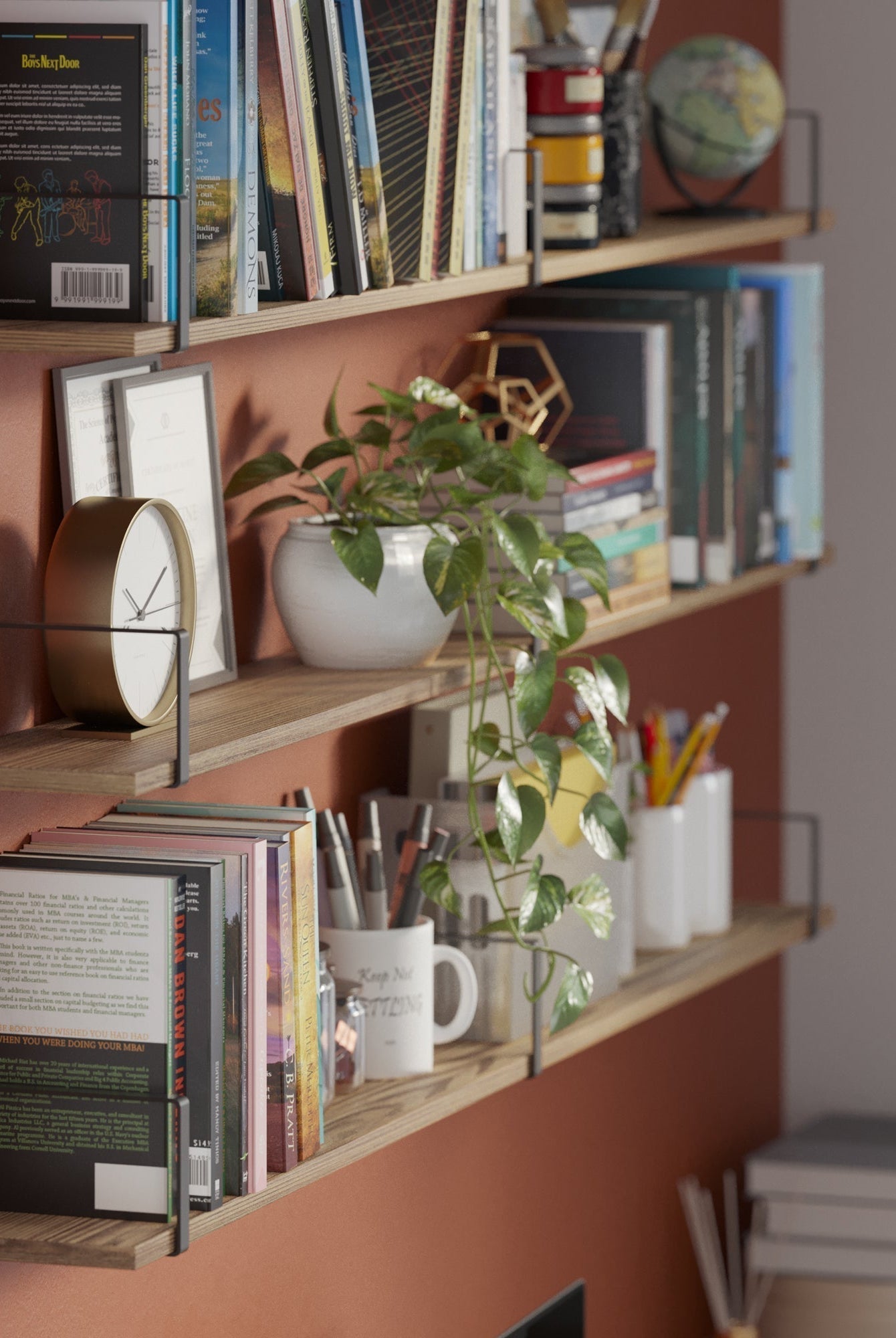 Three wooden wall-mounted shelves with sleek black brackets hold books, stationery, and decorative items against a warm terracotta wall. A trailing green plant adds a lively touch, creating a cozy and functional reading nook.