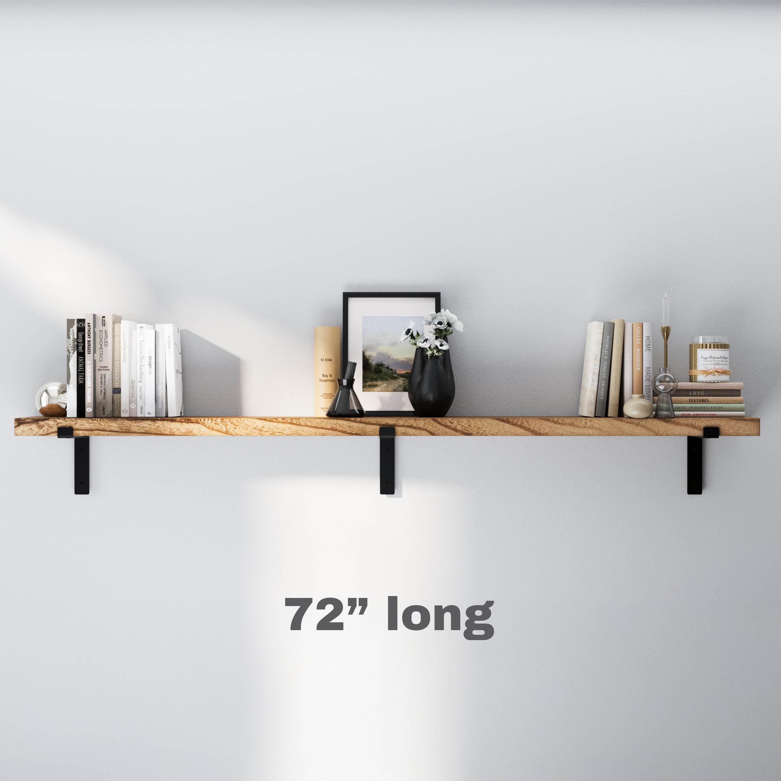 A sleek 72 inch long floating shelf for rustic decor with black brackets, adorned with books, candles, a framed painting, a vase with flowers, and decorative items, creating a balanced and elegant wall-mounted display in a minimalist setting.