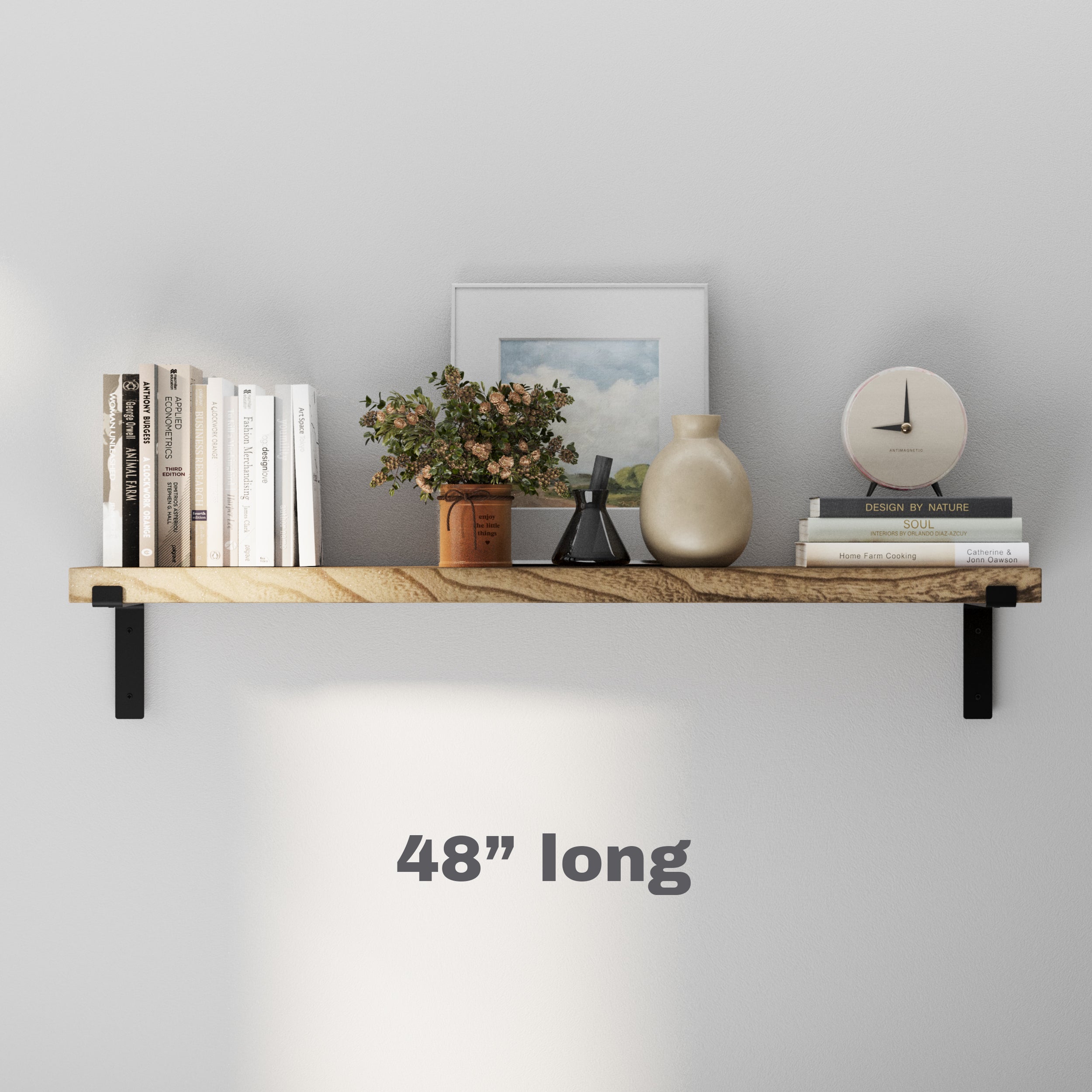 A 48" long wooden display shelf with black brackets, holding books, a clock, decorative vases, and a framed picture against a white wall.