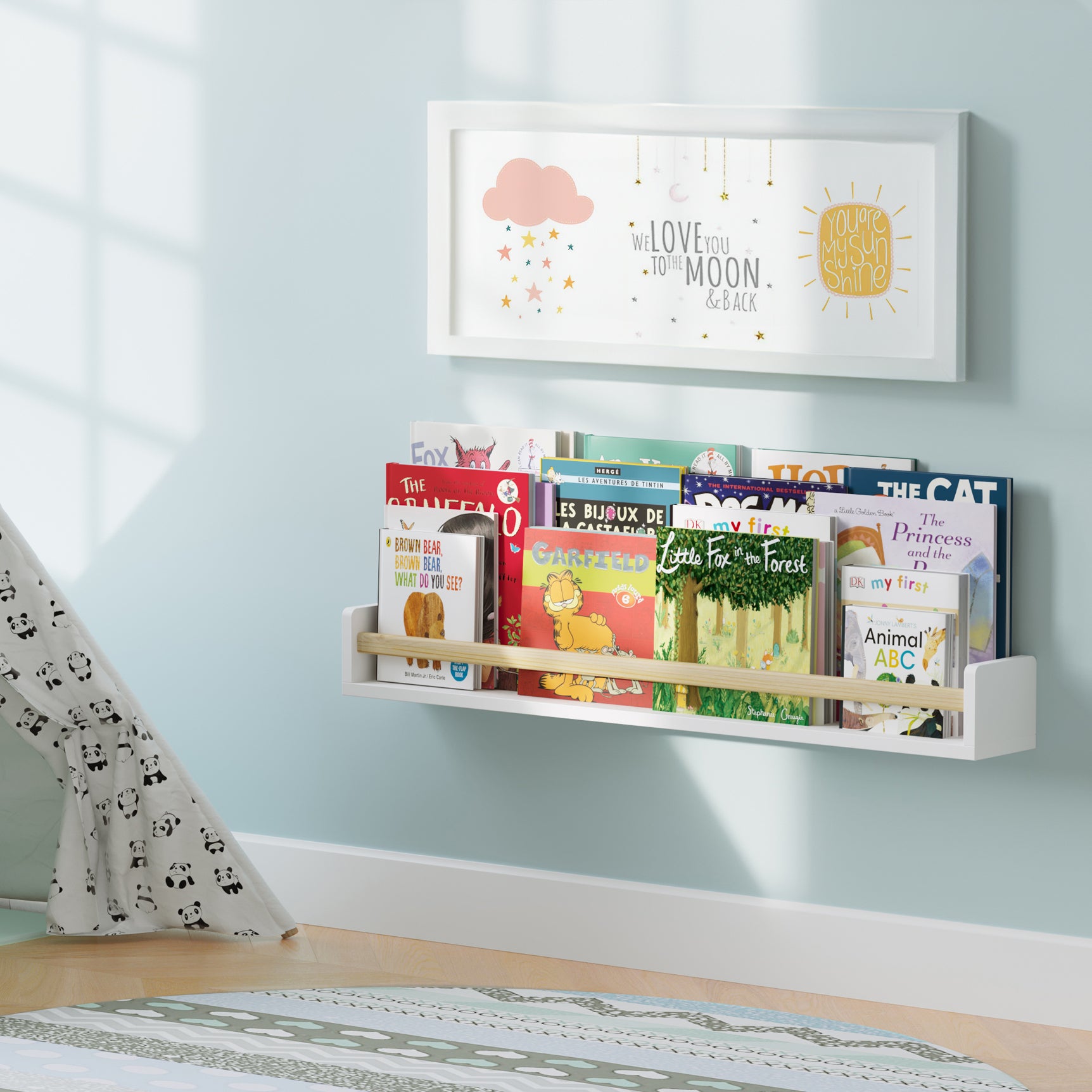 A white nursery book shelf with a natural wood rod in a child's room, filled with children's books. Mounted on a light blue wall near a play tent, creating a cozy reading nook for kids.