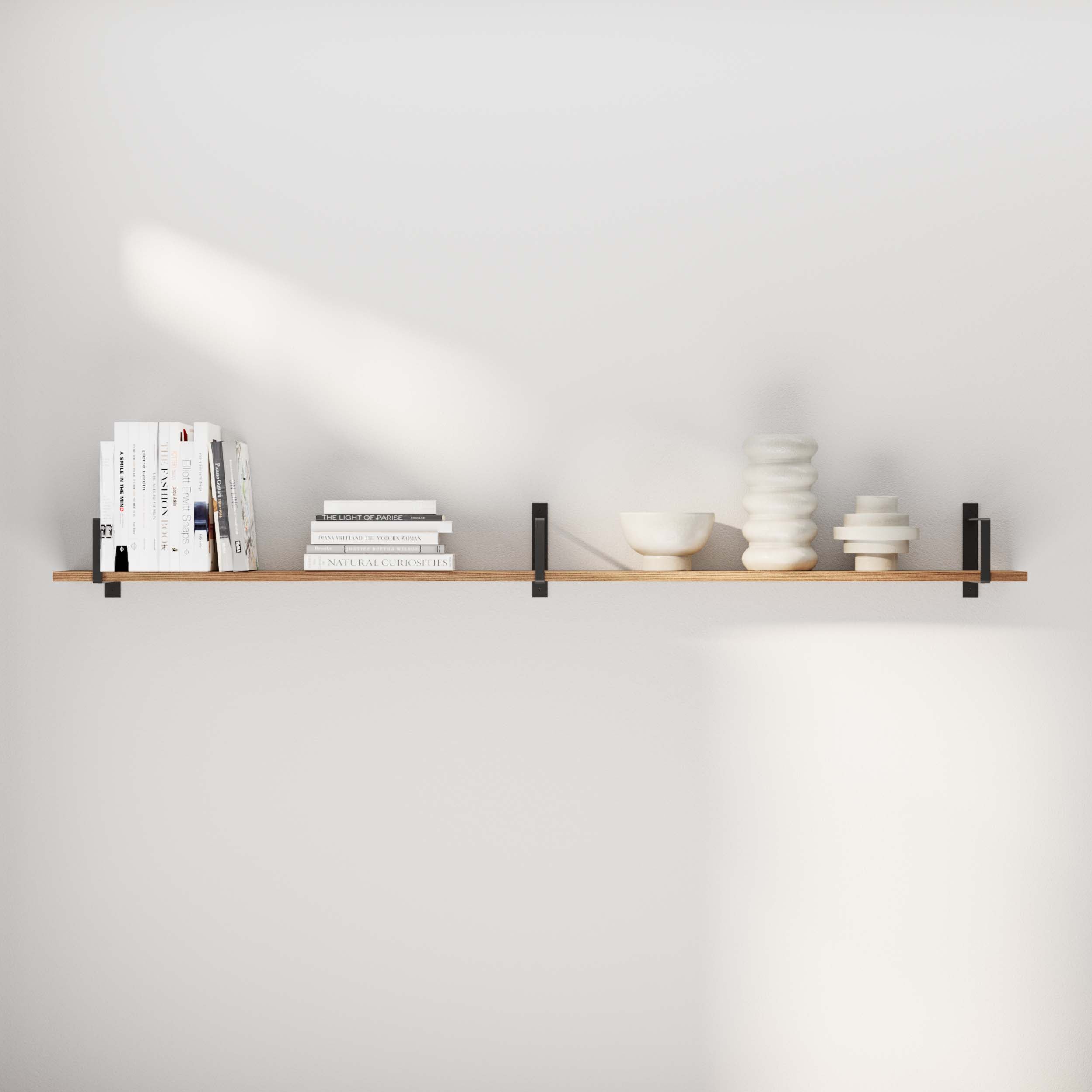 A minimalist and long floating bookshelf showcases neatly stacked white books and modern ceramic décor. Its sleek black metal brackets provide both support and an industrial touch to the clean, neutral-toned room.