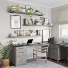 A home office setup with three long wall shelves in rich gray finish displaying books, plants, and decor. Below, a gray desk with a laptop and stationery sits against the wall. A wooden chair and a potted plant complete the cozy workspace.