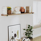 A hanging bookshelf with a warm wood tone, featuring books, vases, and a framed abstract print in a cozy corner.