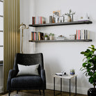 Two wall book shelves in a rich ebobny finish above a dark armchair with books, plants, and modern décor in a reading area featuring a gold floor lamp.