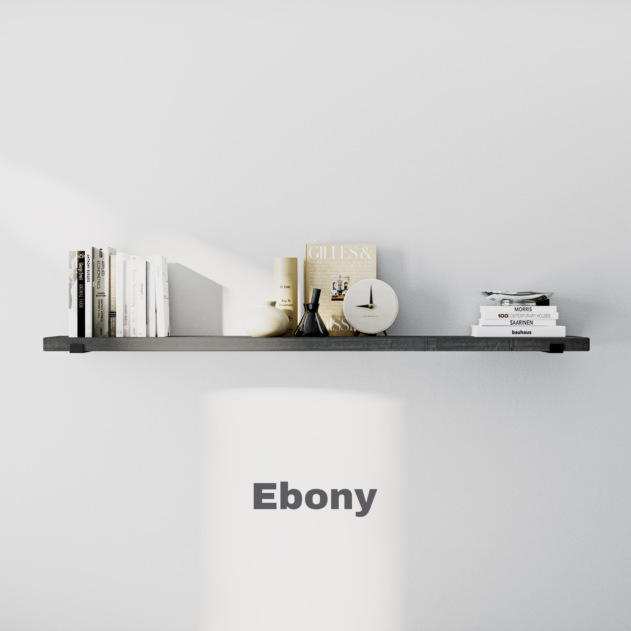 A sleek, ebony-colored 60 inch long floating shelf on a white wall, holding books, a clock, and decorative objects in a minimal style.