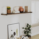 A cozy corner with a rustic hanging shelf in an espresso finish, featuring books, ceramics, and decorative objects. Below, an abstract framed artwork leans against the wall next to a potted plant.