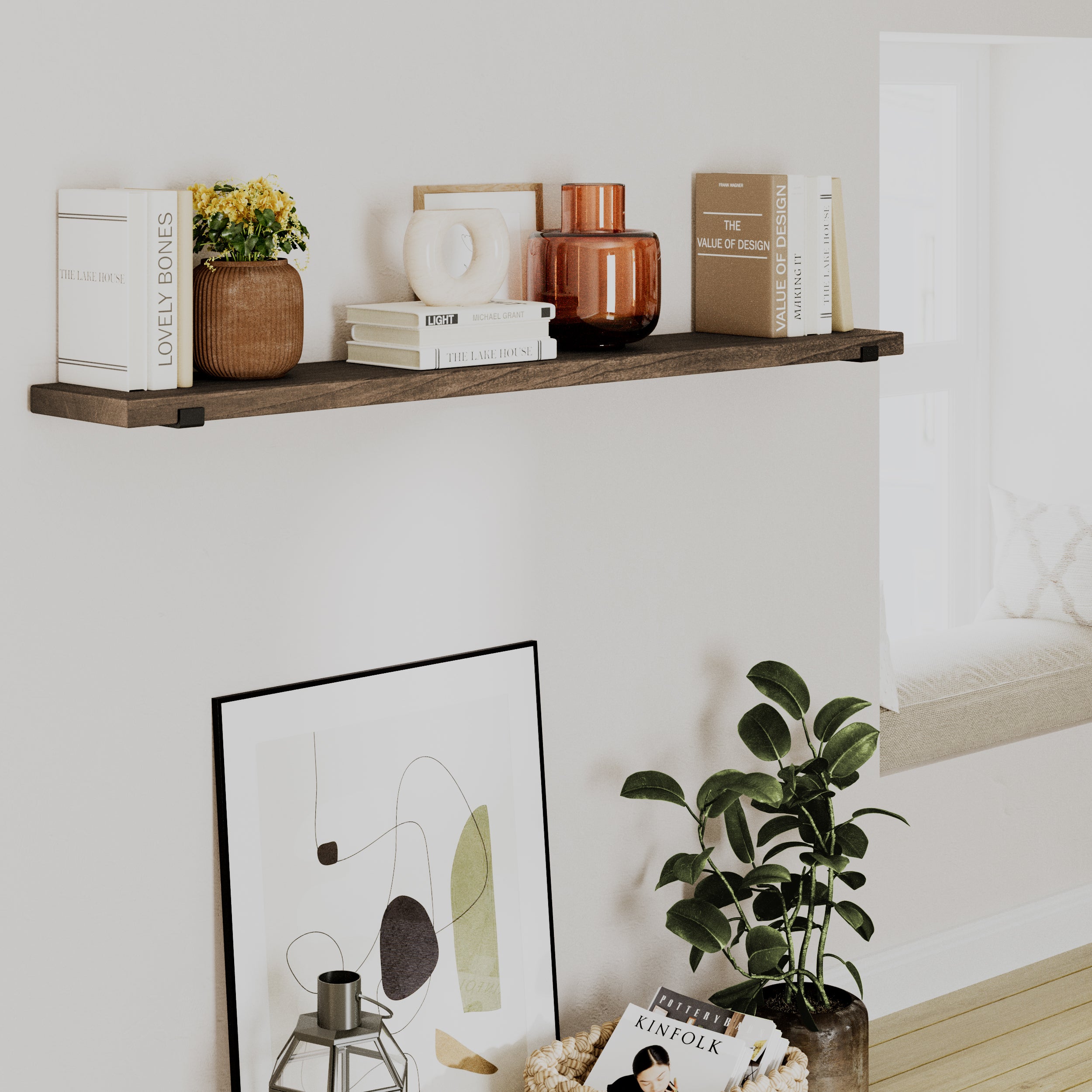 A cozy corner with a rustic hanging shelf in an espresso finish, featuring books, ceramics, and decorative objects. Below, an abstract framed artwork leans against the wall next to a potted plant.