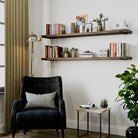 Two espresso-finished long floating shelves hold books, potted plants, and framed art above a black armchair with a gold floor lamp, creating a stylish reading area.