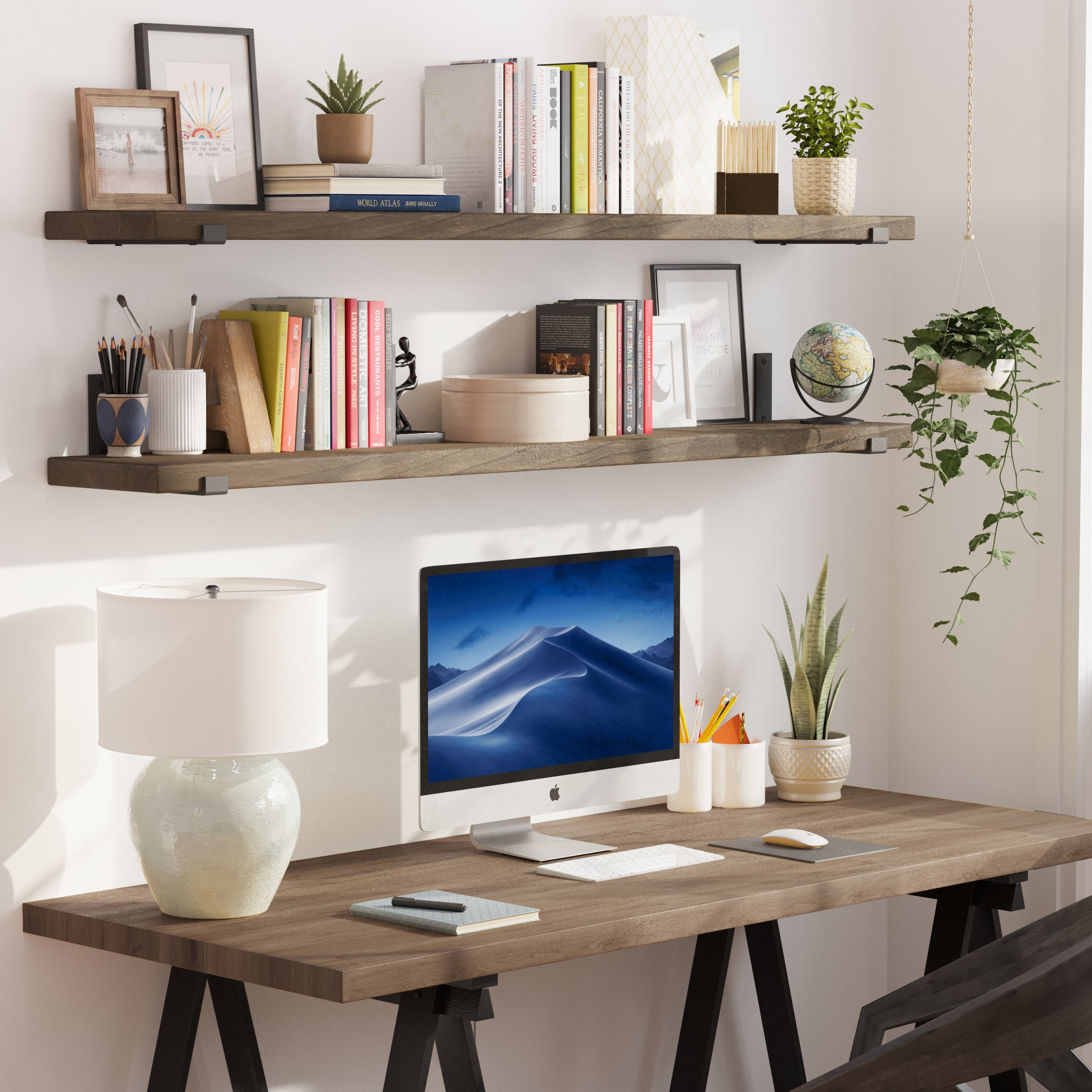 Two espresso-finished floating office shelves above a wooden desk display books, plants, and office essentials. A Mac computer and a desk lamp complete the modern workspace.