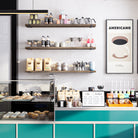 60 inch long floating shelves in espresso color hold coffee essentials, bags of beans, and decorative items above a counter with a menu board, pastries, and syrup dispensers, giving a chic café vibe.