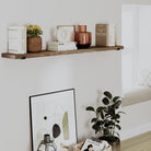 A single provincial wood shelf above a seating area, decorated with books, vases, and framed artwork. A green plant adds natural contrast.