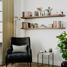 Two long wooden shelves in provincial finish mounted above a cozy reading nook with a dark armchair, books, and plants.