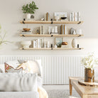 Three floating shelves in Natural finish hold books and ceramics above a white wainscoted wall, complementing a cozy and bright living room with neutral tones.