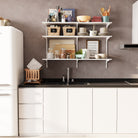 A minimalist kitchen scene featuring a 3-tier white wall-mounted kitchen shelf unit. It stores tea and coffee baskets, plates, cups, jars, cookbooks, and decor—blending function and design beautifully above a modern sink.