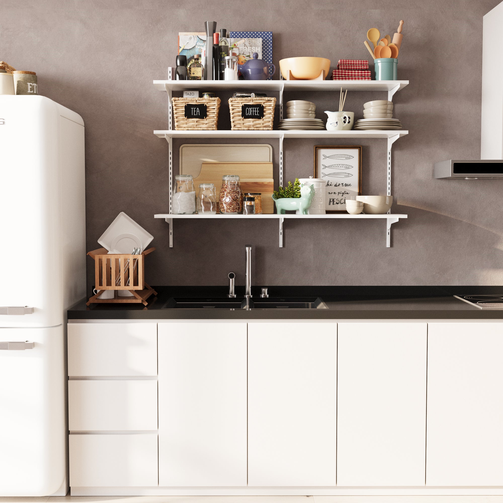 A minimalist kitchen scene featuring a 3-tier white wall-mounted kitchen shelf unit. It stores tea and coffee baskets, plates, cups, jars, cookbooks, and decor—blending function and design beautifully above a modern sink.