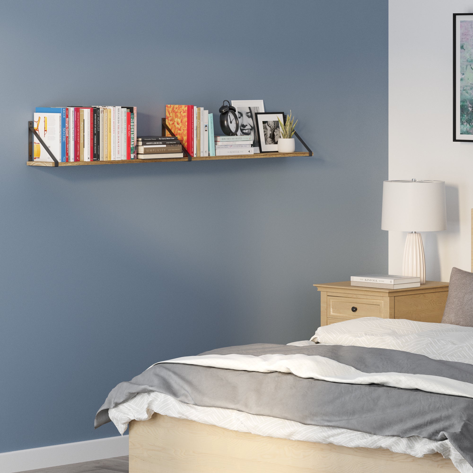 A part of a bedroom with a blue wall. There's a book shelf for bedroom above the bed filled with books and some framed pictures. Beside the bed is a wooden nightstand with a lamp on it, and the bed itself is partially visible with a grey and white bedding.