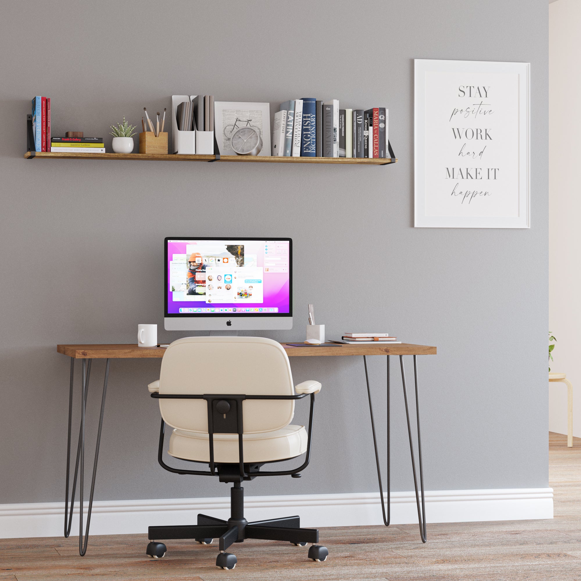 Against a gray wall is a wooden desk holding an all-in-one computer, with a mug and some notes on the side. Above the desk, there's a wall book shelf filled with books, organizers, and a small plant, contributing to a functional and stylish look. To the right on the wall, there's an inspirational framed poster with the text "STAY positive WORK hard MAKE IT happen."