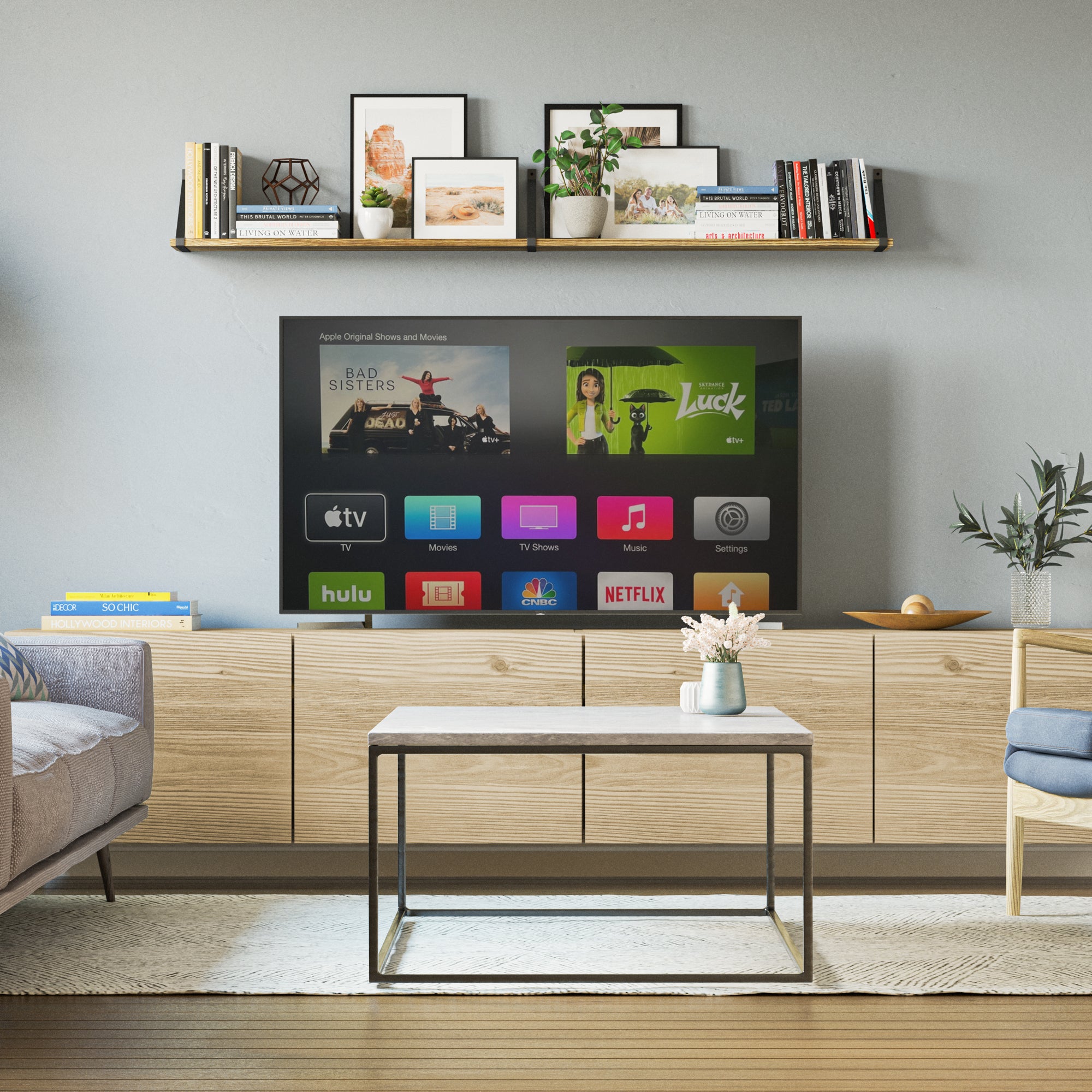 A modern living room with a flat-screen TV displaying a streaming service interface on a gray wall. Above the TV, there is a long shelf with books, framed pictures, and plants. Below the TV, a wooden media console stretches across, with a small coffee table and decorative items in the foreground.