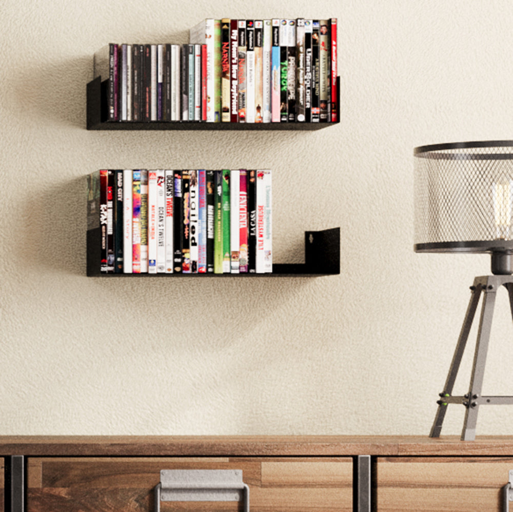 Two metal floating shelves in a rich black finish mounted on a beige wall hold an organized collection of DVDs and books. Below, a wooden console with metal accents provides storage. To the right, a modern tripod lamp with a mesh shade adds style.