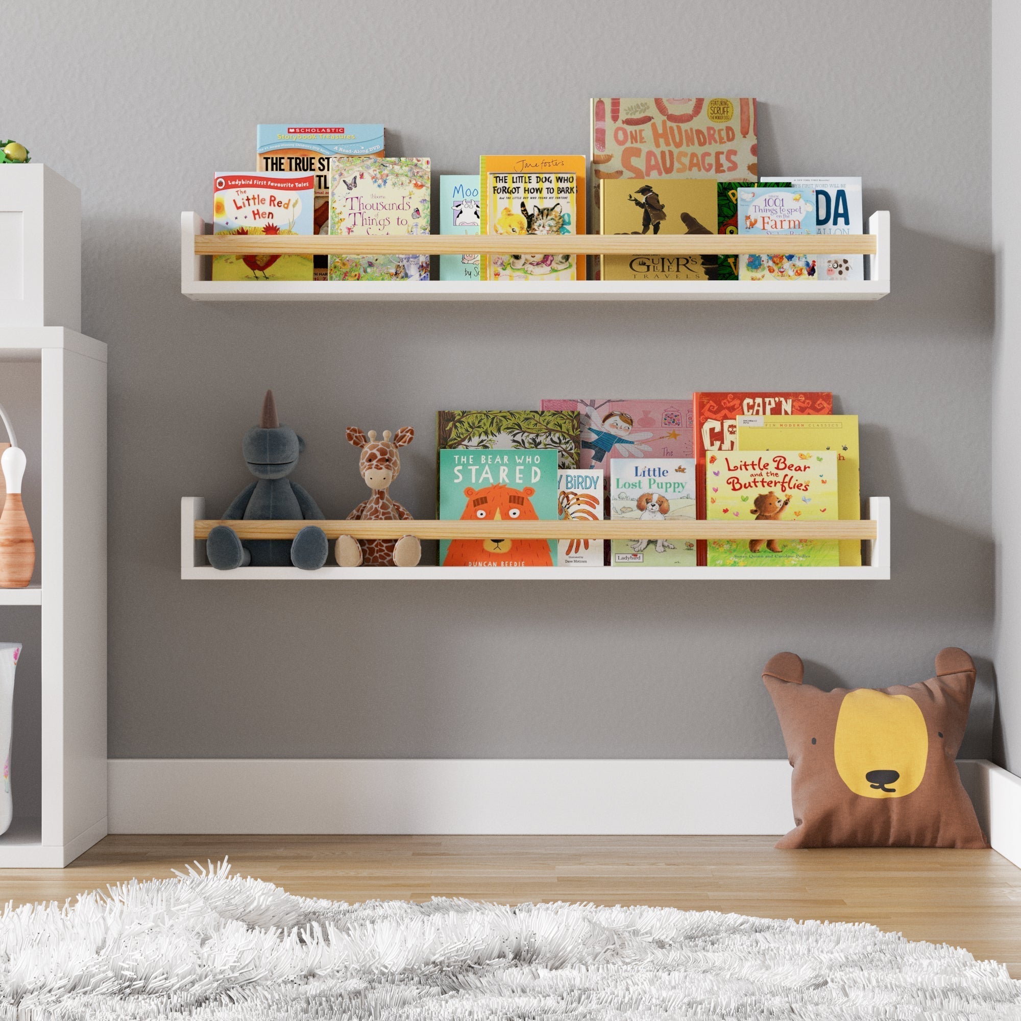 A cozy children's reading nook with two white wall-mounted bookshelves filled with colorful picture books. A plush bear pillow rests on the floor beside a fluffy rug, with a small stuffed animal sitting on the lower shelf.