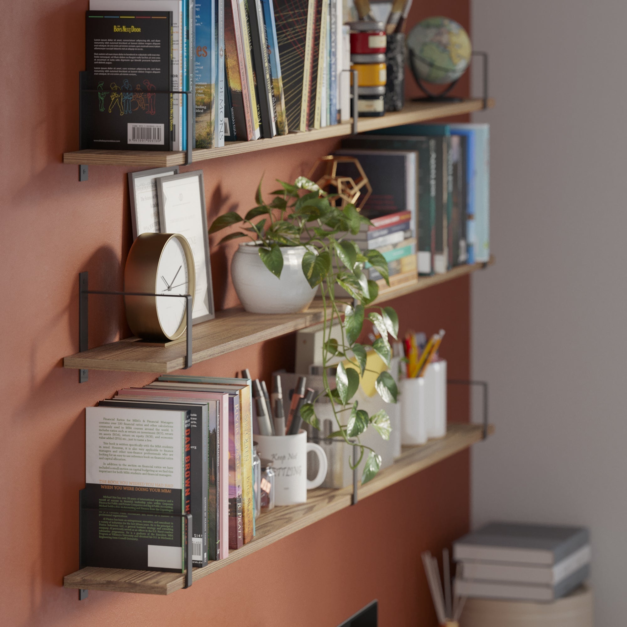 Three wooden wall-mounted shelves with sleek black brackets hold books, stationery, and decorative items against a warm terracotta wall. A trailing green plant adds a lively touch, creating a cozy and functional reading nook.