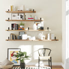 A well-lit wall featuring multiple wooden rustic shelves with different length options, each adorned with books, vases, candles, and decor items. A black chair, green plant, and framed artwork below add charm, creating a cozy and stylish living space.