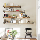 A stylish staggered shelf layout with five rustic wood shelves mounted with black metal brackets. Displayed with art, books, vases, and minimal decor, creating an eye-catching accent wall above a black chair.