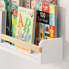 A close-up of the wooden nursery shelf white holding children's books, emphasizing the sturdy wooden rod and sleek white frame. The design is practical and stylish, ideal for organizing and displaying books.