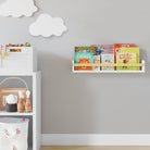 A floating nursery bookshelf holding children's books, mounted on a light gray wall with white cloud decorations. Positioned above a white shelf unit with wooden toys, creating a charming and tidy kids' space.