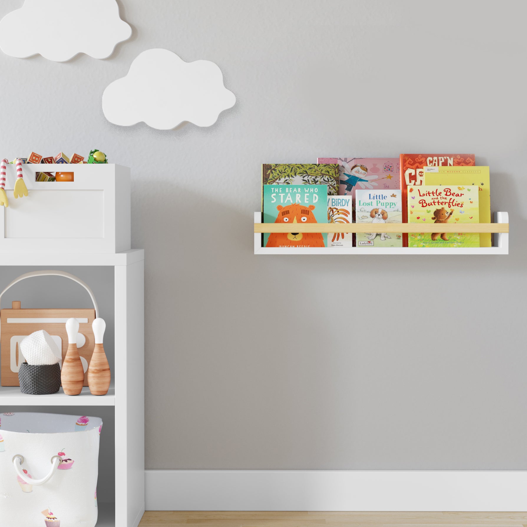 A floating nursery bookshelf holding children's books, mounted on a light gray wall with white cloud decorations. Positioned above a white shelf unit with wooden toys, creating a charming and tidy kids' space.