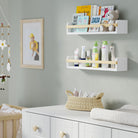 Two white shelves for nursery in a nursery, one holding children's books and the other baby care products. Mounted above a white dresser with a woven basket, creating an organized and cozy space for a baby.