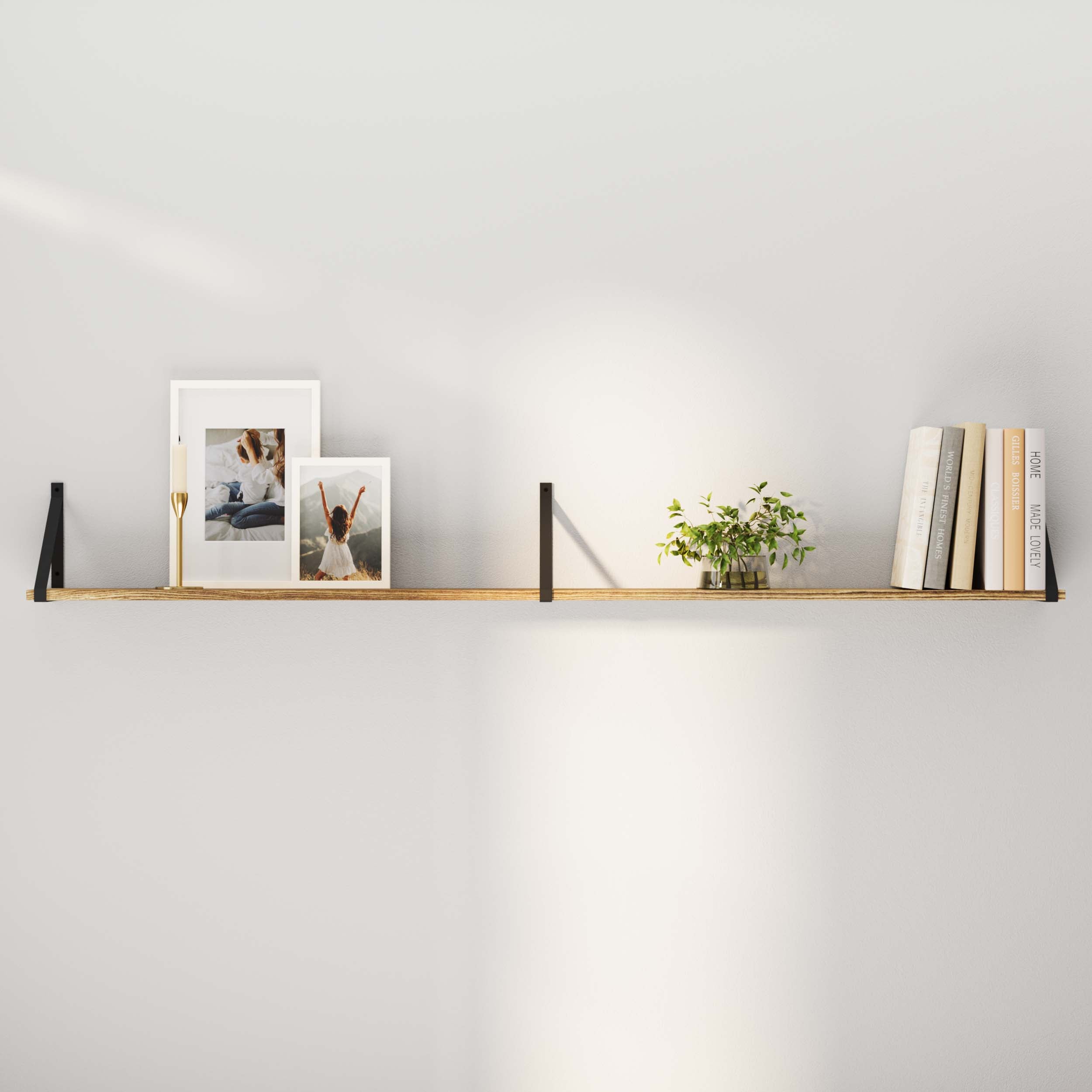 A burnt floating shelf against a white wall, featuring two framed photographs, a small potted plant, and a row of books.