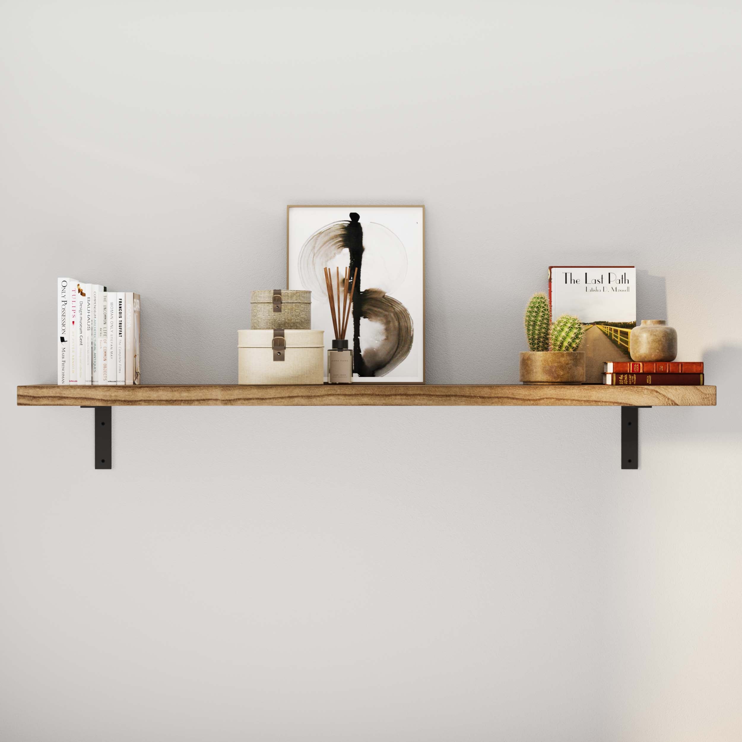 A rustic shelf affixed to a light-colored wall by two sturdy metal brackets with books, decorative boxes, a framed artwork, a cactus, and candles.