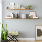 A modern setup featuring two floating wooden shelves 48" and 60" length against a light blue wall. Decorated with books, vases, plants, and framed art, it exudes an elegant, minimalist style. Below, a green cushioned bench and potted plant add a cozy touch.