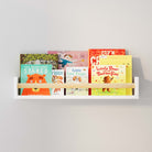 A white nursery book shelf with a natural wood rod filled with children's books, mounted on a light gray wall. The shelf holds colorful book covers, creating an inviting and organized space for young readers.