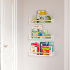 Three nursery book shelves with natural wood rods holding children's books and toys, mounted on a light pink wall beside a doorway. The setup creates an organized and charming reading nook for kids.
