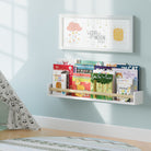 A white nursery book shelf with a natural wood rod in a child's room, filled with children's books. Mounted on a light blue wall near a play tent, creating a cozy reading nook for kids.