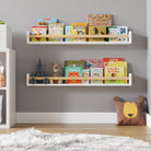 Two white floating book shelves for wall nursery with natural wood rods filled with children's books and toys, mounted on a light gray wall. Positioned beside a white storage unit and a plush bear pillow, creating a cozy and organized reading space.