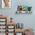 A white display shelf for kids with a natural wood rod in a child's room, holding large books. Mounted on a light blue wall above a wooden toy storage unit, creating a colorful and organized reading area.