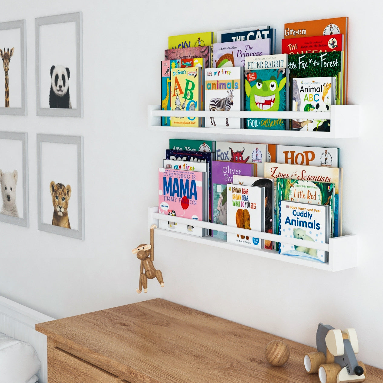 A nursery with two white wooden shelves for kids filled with colorful books above a wooden dresser, decorated with animal-themed wall art and a hanging monkey toy.