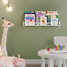 A nursery room with a white floating shelf for kids room decor on a green wall, holding books, next to a giraffe plush toy and a small table with art supplies.
