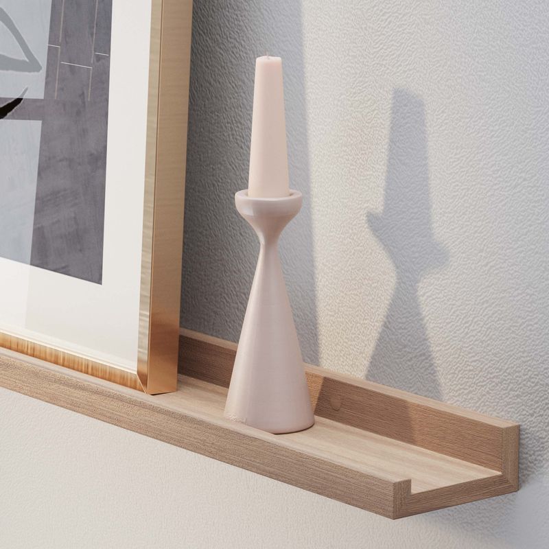 A long wall shelf with a pale pink candlestick is in focus, next to a framed abstract artwork, against a textured wall.