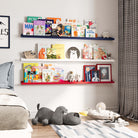 a children's room with three shelves mounted on the wall, showcasing a variety of colorful books. The shelves are color-coded, with the top shelf in blue, the middle in white, and the bottom in red. Below the shelves, there is a soft toy shaped like a dog lying on a striped rug. The room's overall design is playful and cozy, with elements like a clock, framed pictures, and stuffed animals enhancing the space.