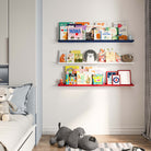 three wall-mounted shelves in different colors (blue, white, red) filled with children's books in a child's room. A plush dog toy is on the floor, creating a cozy, organized reading space.