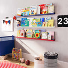 Cute nursery shelves are shown in a different children's room setting. The color scheme includes a blue shelf at the top, a white shelf in the middle, and a red shelf at the bottom, consistent with the previous images. The room has a playful vibe, with a variety of books, a toy figure of Superman, and a colorful wall decoration. A small wooden toy, a laundry basket, and a football add to the child-friendly atmosphere.