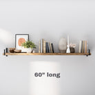 A 60" long floating bookshelf features a solid wood board supported by metal side brackets. It holds a mix of books, decorative vases, greenery, and framed art, styled against a soft gray wall with a minimalist look.