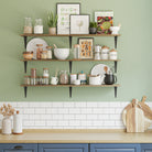 Triple kitchen shelves on a green kitchen wall, stocked with jars, herbs, cookbooks, and kitchenware—perfect for storage and rustic kitchen decor.
