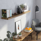 A single wood floating shelf for wall decor showcases a mix of books, glass bottles, and greenery against a soft gray wall. Styled with a minimalist touch, it creates a serene corner ideal for relaxing with a warm, earthy aesthetic.
